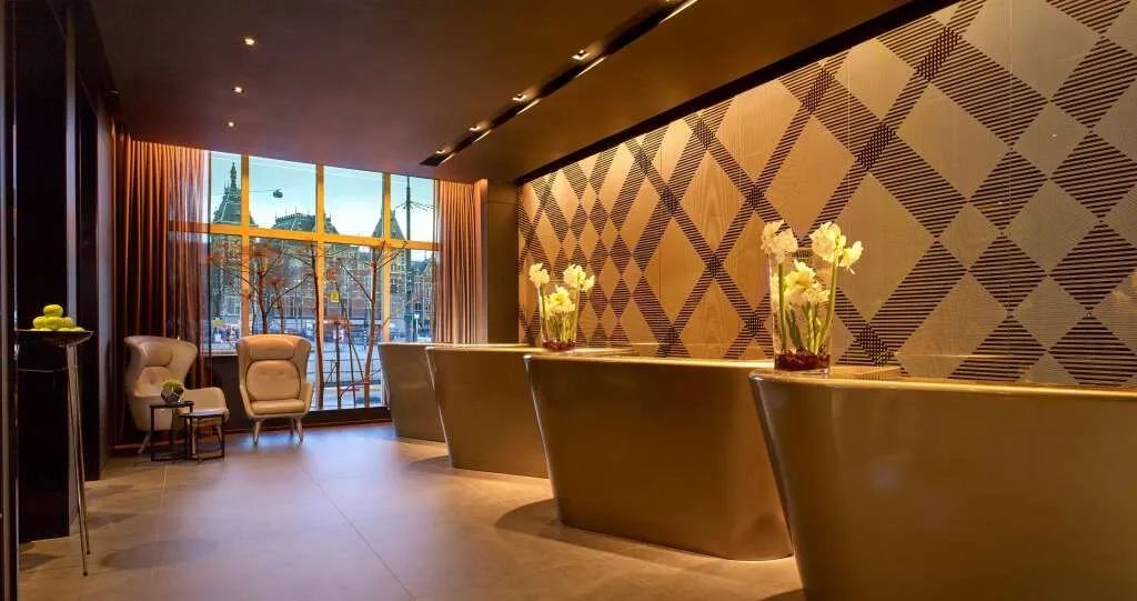 An interior shot of a modern reception area, likely a hotel lobby, featuring several stylish reception desks with floral arrangements, a geometrically patterned wall, and a large window overlooking an urban streetscape.