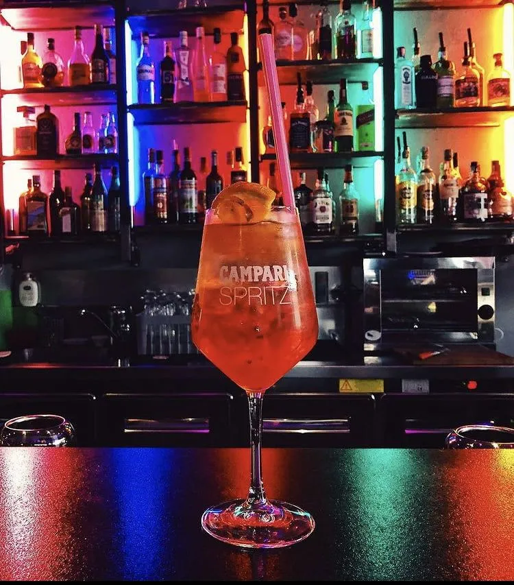 The image features a Campari Spritz cocktail in a large stemmed glass placed on a bar counter. The background shows shelves filled with various liquor bottles, illuminated by colorful, vibrant lights. The composition highlights the drink as the central subject, with reflections of the colored lights visible on the bar surface.