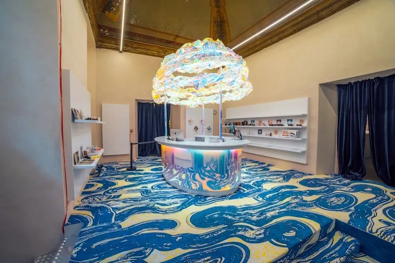 The image features a brightly lit and modern interior space, likely a retail store or gallery, dominated by a circular counter with an iridescent base and a large, ornate, cloud-like light fixture hanging above it. The floor is covered with a vibrant blue and yellow abstract patterned carpet that resembles water or waves. Shelves displaying various items are visible on the walls, and the overall aesthetic is artistic and contemporary.