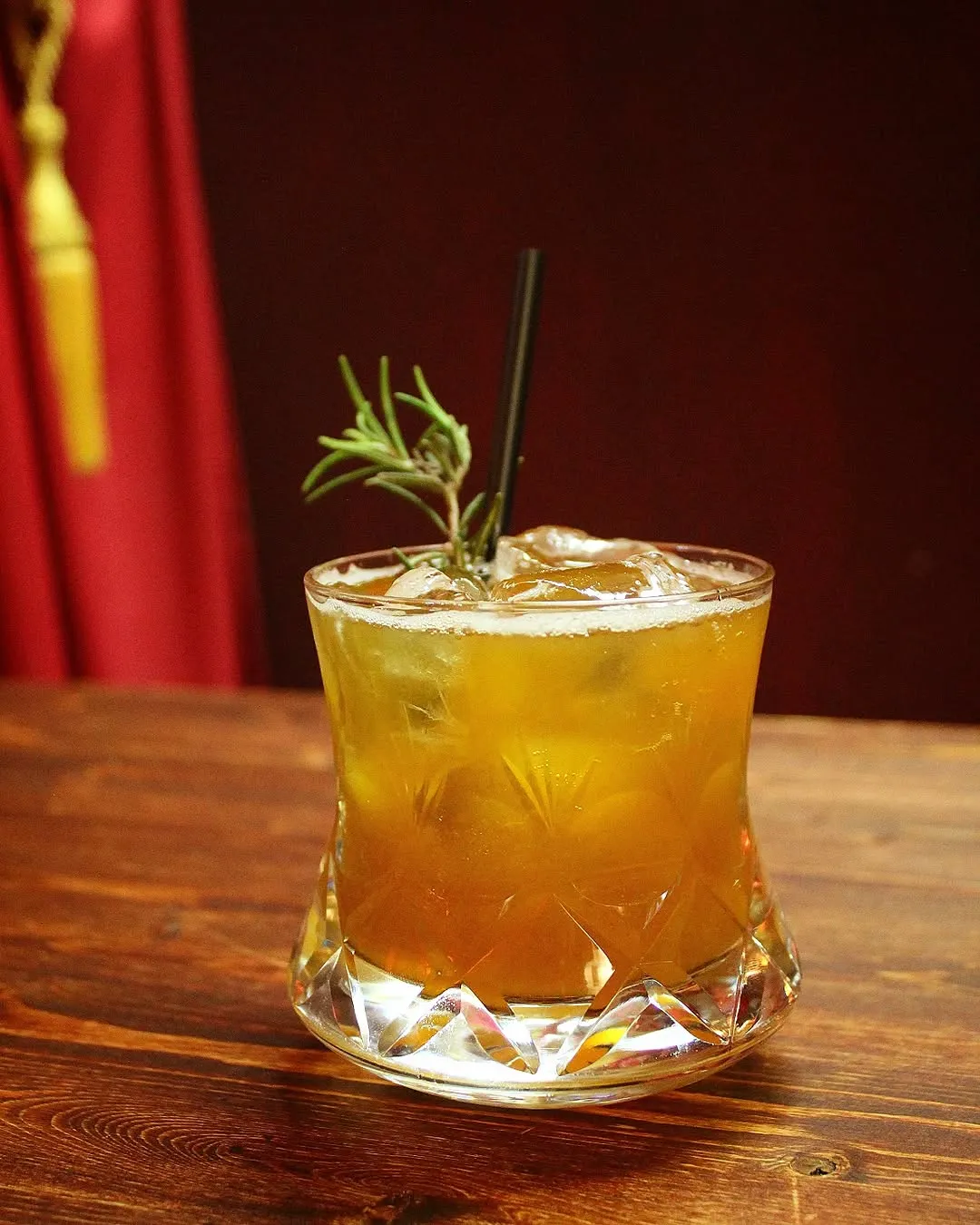 A close-up shot of a cocktail in a cut glass, garnished with rosemary and ice, resting on a wooden table, with a red curtain in the background.