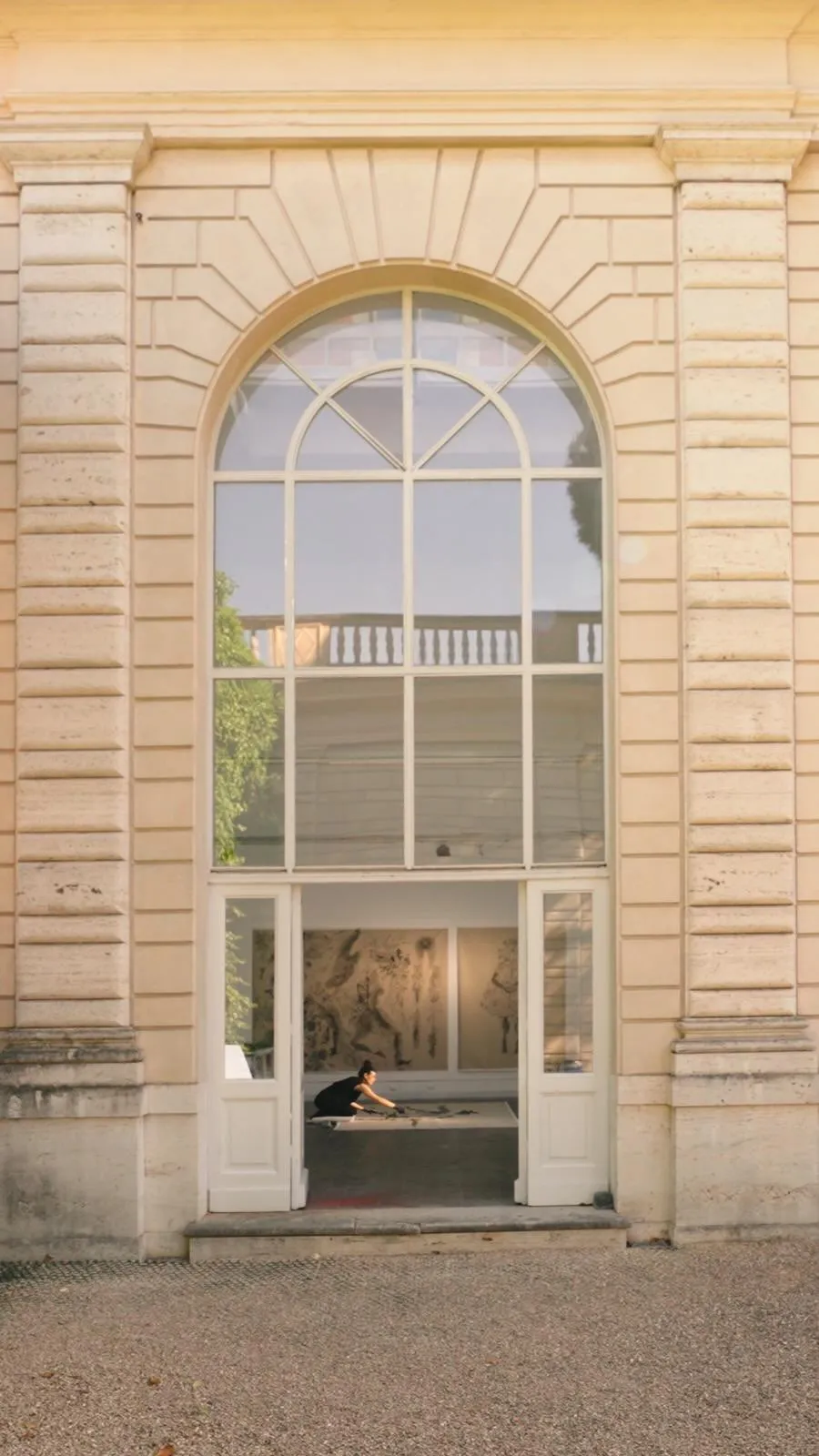 An external view of a grand building facade featuring a large, arched multi-paned window, through which a person is visible working inside what appears to be an art studio. The building is constructed from light-colored stone.