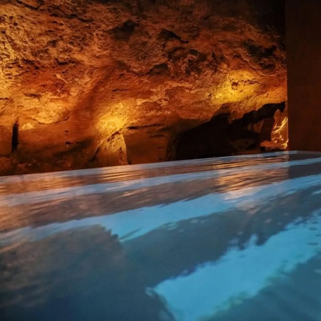 The image is well-composed, capturing an atmospheric and inviting spa environment. The moody lighting effectively highlights the textured rock formations and the reflective water surface, contributing to a strong sense of depth and tranquility. The photo is highly photo-realistic with good color accuracy.