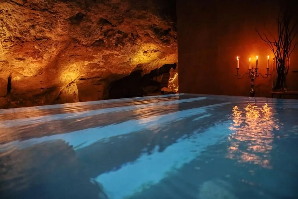 The image is well-composed, capturing an atmospheric and inviting spa environment. The moody lighting effectively highlights the textured rock formations and the reflective water surface, contributing to a strong sense of depth and tranquility. The photo is highly photo-realistic with good color accuracy.