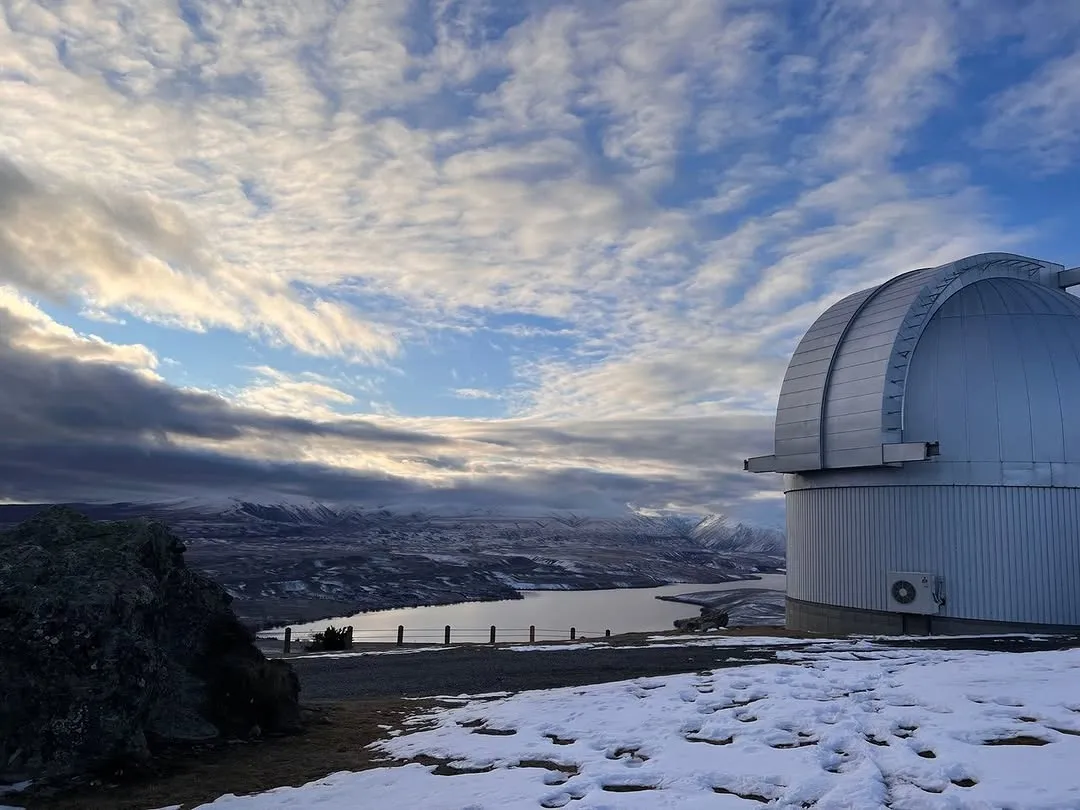 An image of an astronomical observatory dome situated on a snow-covered hill, overlooking a lake and distant mountains under a cloudy sky.