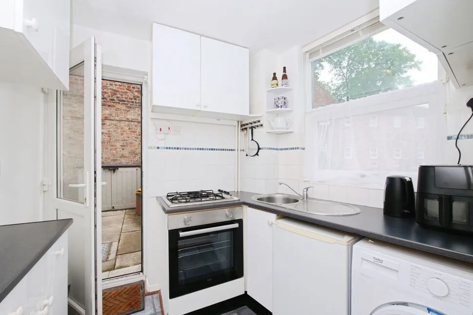 A well-lit and photo-realistic image of a small, functional kitchen. The key elements, such as the stove, oven, and sink, are sharp and in focus. The colors appear accurate, and the composition effectively captures the room's layout, including a view through an open door.
