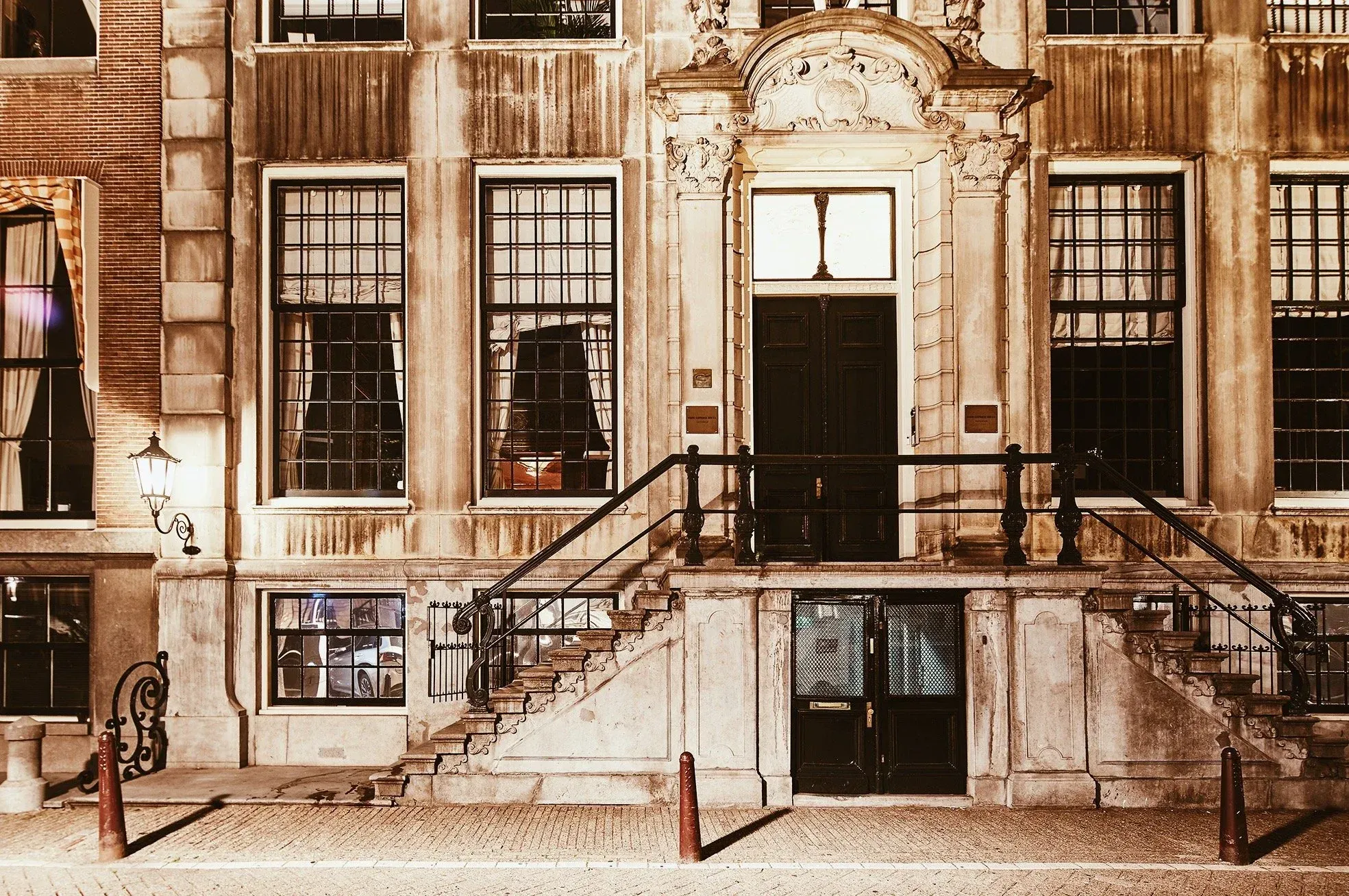 The image features a well-composed shot of an old building facade, exhibiting good sharpness and photo-realism. The architectural details, including the ornate entrance and stone staircase, are clearly in focus. The artificial lighting creates a distinct warm, almost golden-hour like aesthetic, contributing to an editorial style. However, some areas in the windows appear overexposed due to interior lighting, and the overall color accuracy might be influenced by the chosen warm tone.