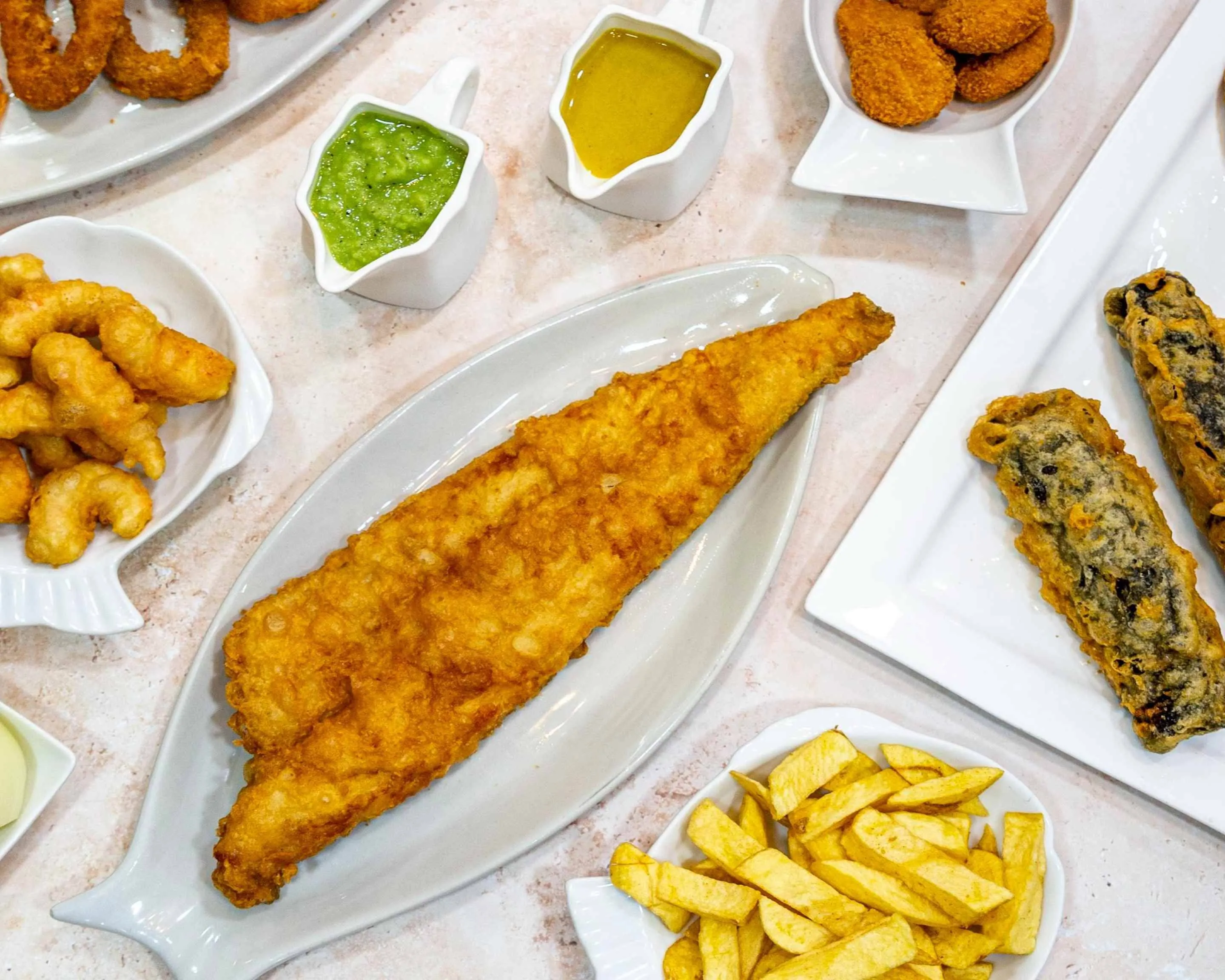 A top-down view of a classic fish and chips meal, featuring a large, golden-brown fried fish fillet on a white platter, accompanied by a serving of golden fries, and two small white gravy boats containing green mushy peas and yellow curry sauce, all arranged on a light-colored, textured surface.