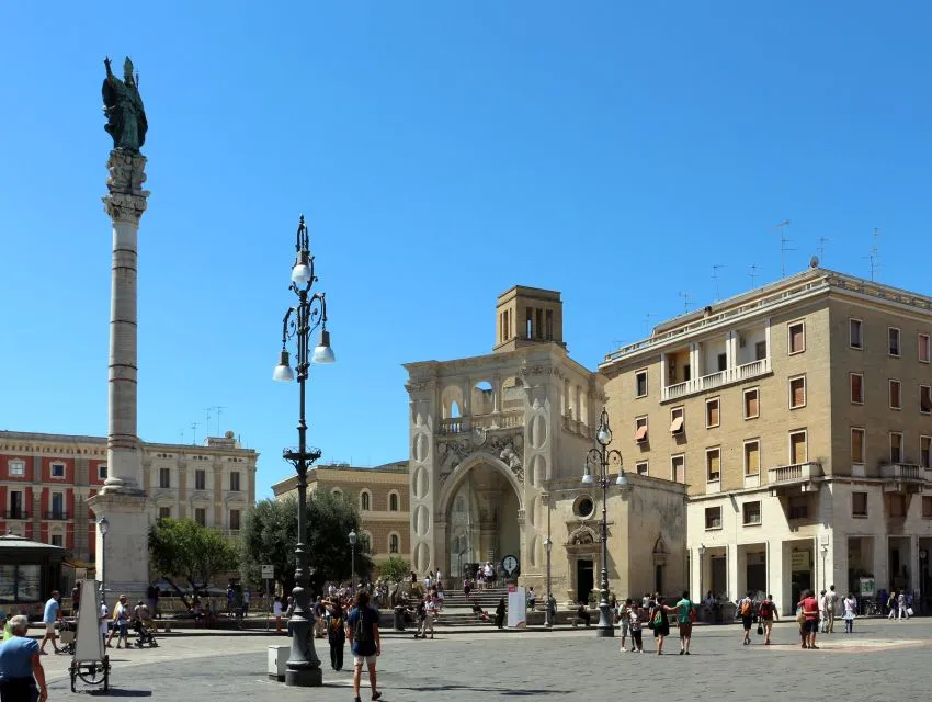 The image features a sunny European city square with prominent historical architecture, including a large decorative archway and surrounding buildings. People are visible strolling through the square, adding a sense of activity. The sky is clear blue.