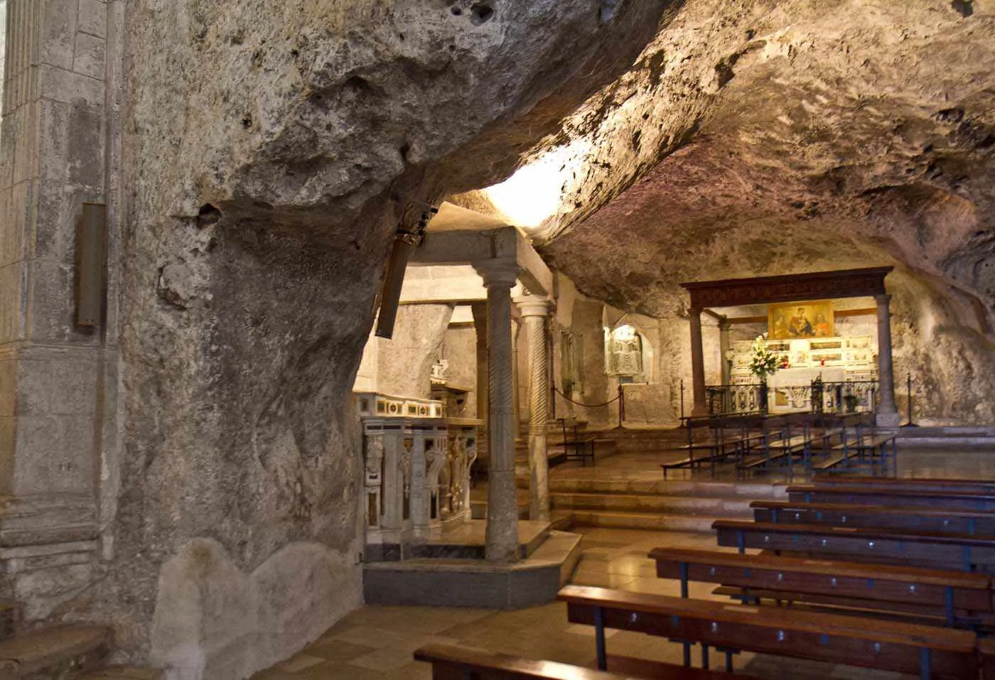 An interior view of a church built within a natural cave, featuring carved stone elements, wooden pews, and an altar area.
