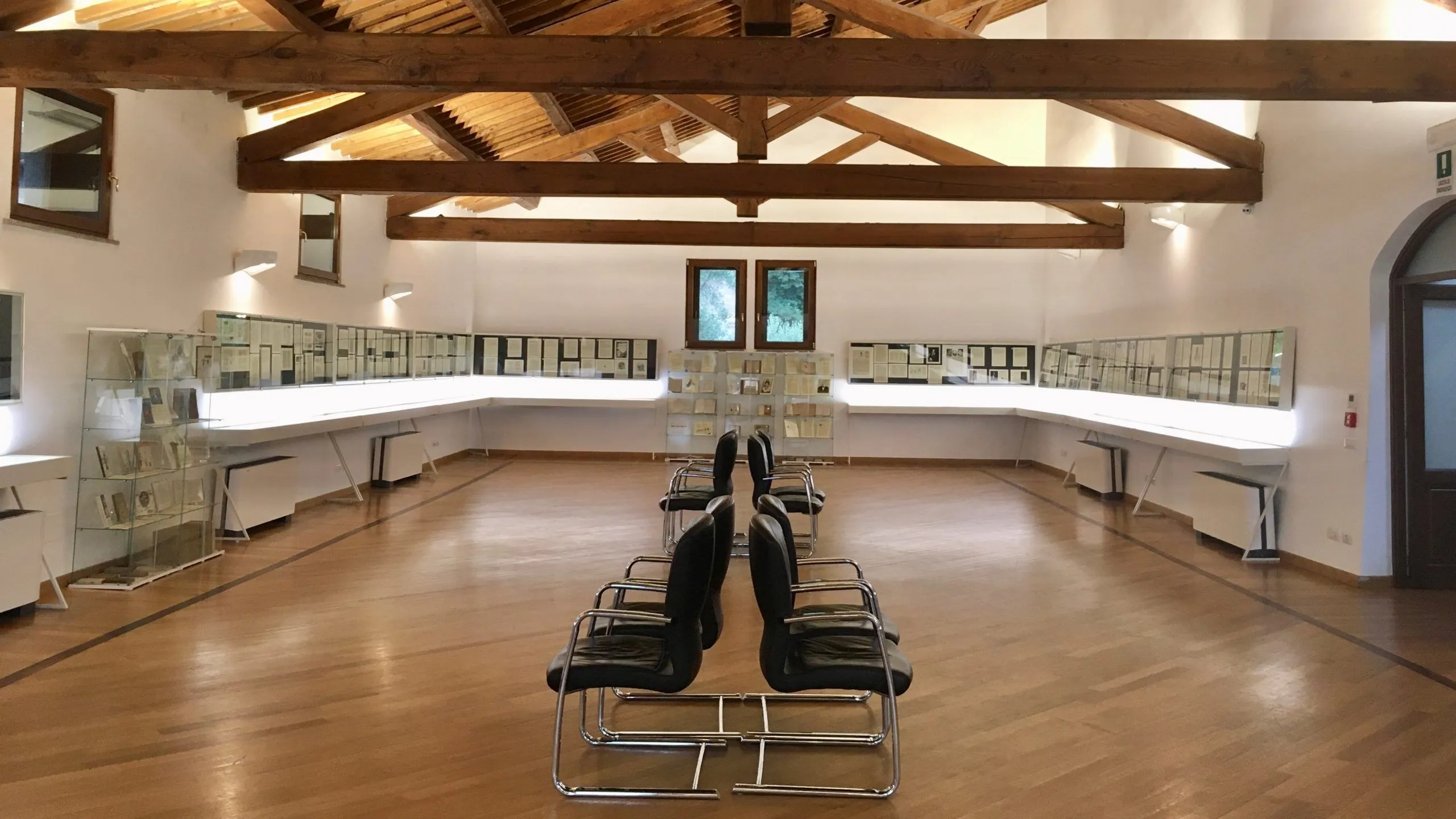 The image displays an exhibition hall or museum interior with rows of chairs facing display cases. The space features a high ceiling with exposed wooden beams and large windows at the far end.