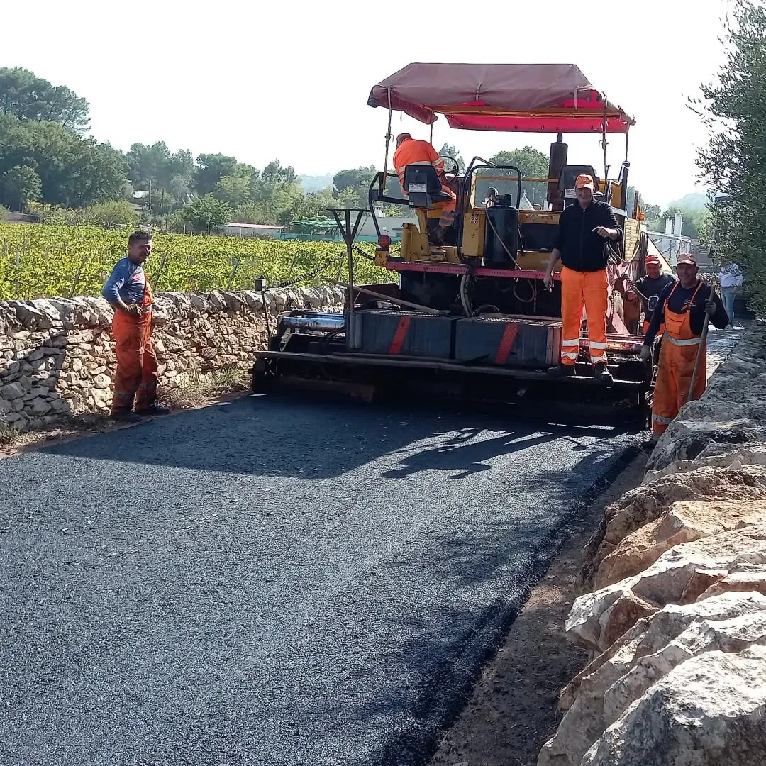 The image effectively captures a road paving operation, demonstrating photo-realism and clear focus on the key elements like the paving machine, workers, and newly laid asphalt. The colors are natural, and the image is generally sharp.
