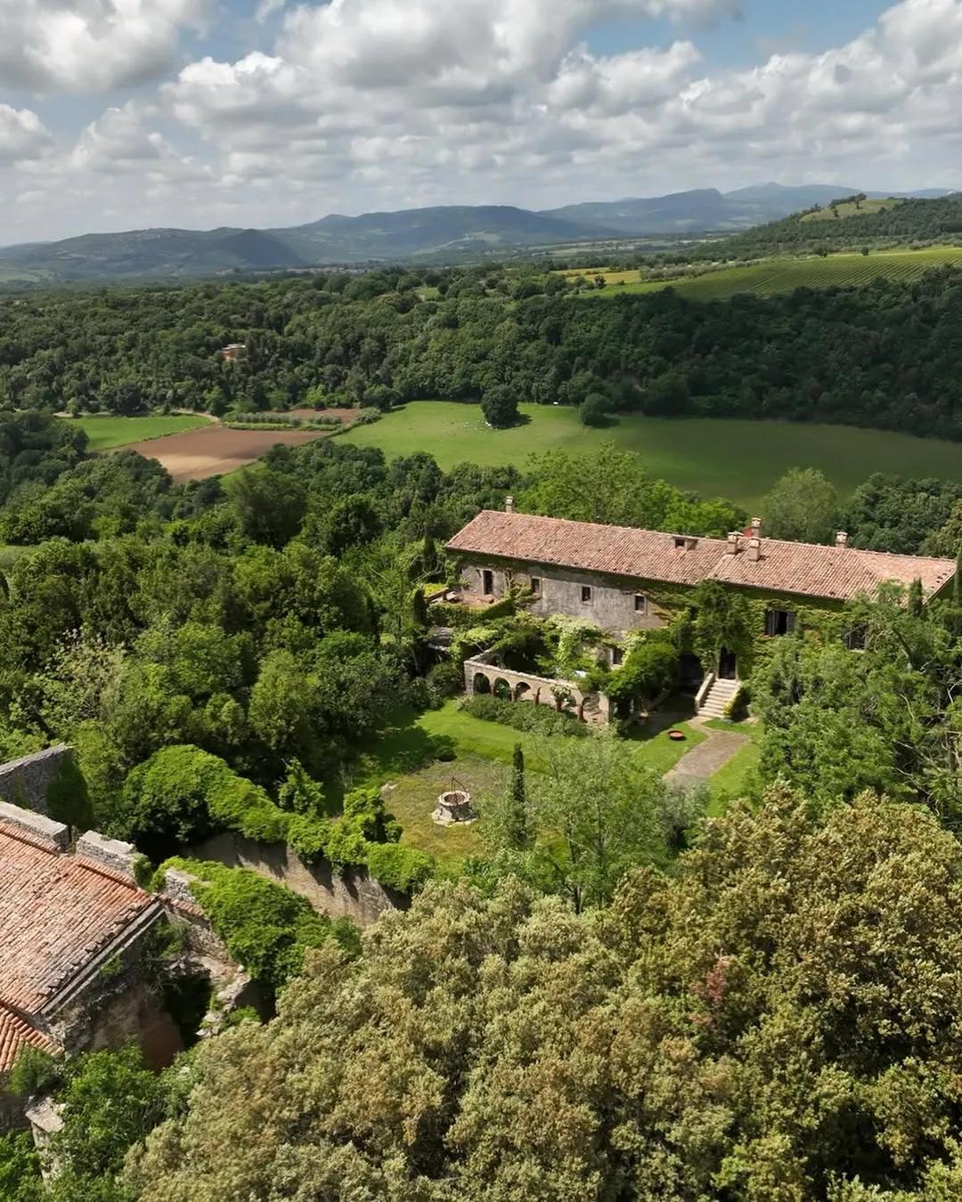 An aerial view of a traditional stone house with a tiled roof, nestled amidst lush green forests and rolling hills, under a partly cloudy sky. Distant mountains form the background.