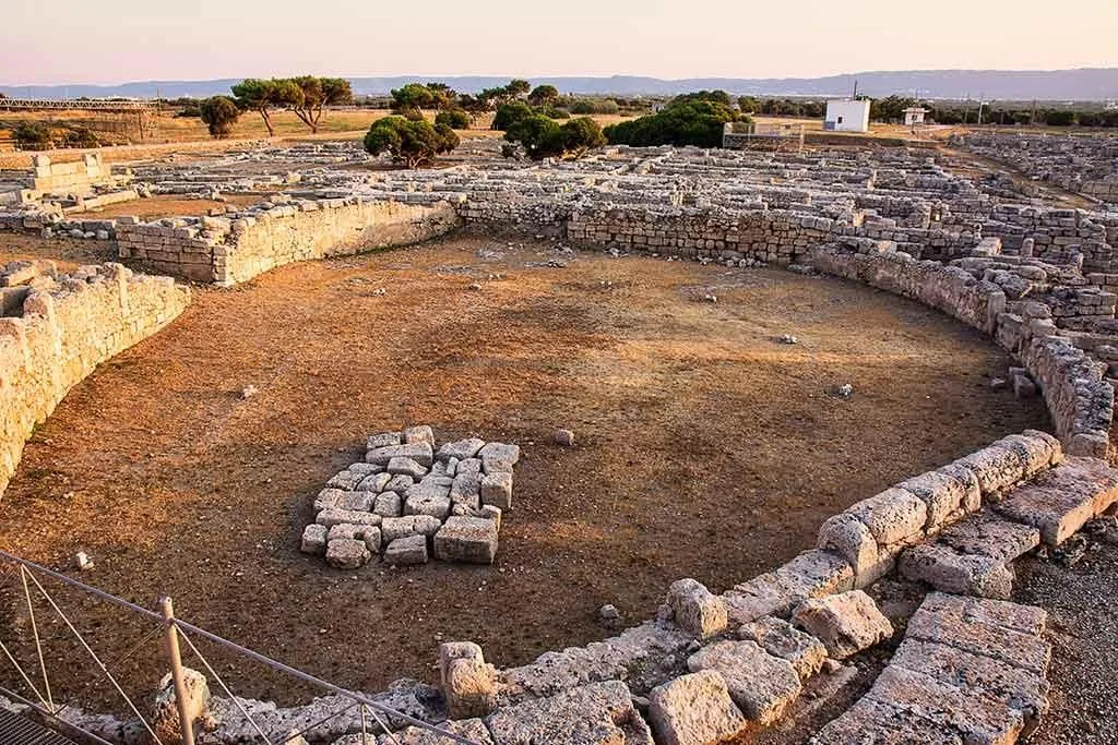 An aerial view capturing the intricate layout of ancient stone ruins at an archaeological site. A prominent circular stone structure dominates the foreground, surrounded by other excavated foundations and remnants of past civilizations. The dry, earthy ground fills the central area, with scattered rocks and distant trees and a small building under a clear sky.