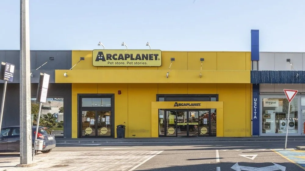 An exterior view of a bright yellow Arcaplanet pet store building, featuring its prominent sign and two entrances, set against a clear blue sky and a parking lot foreground.