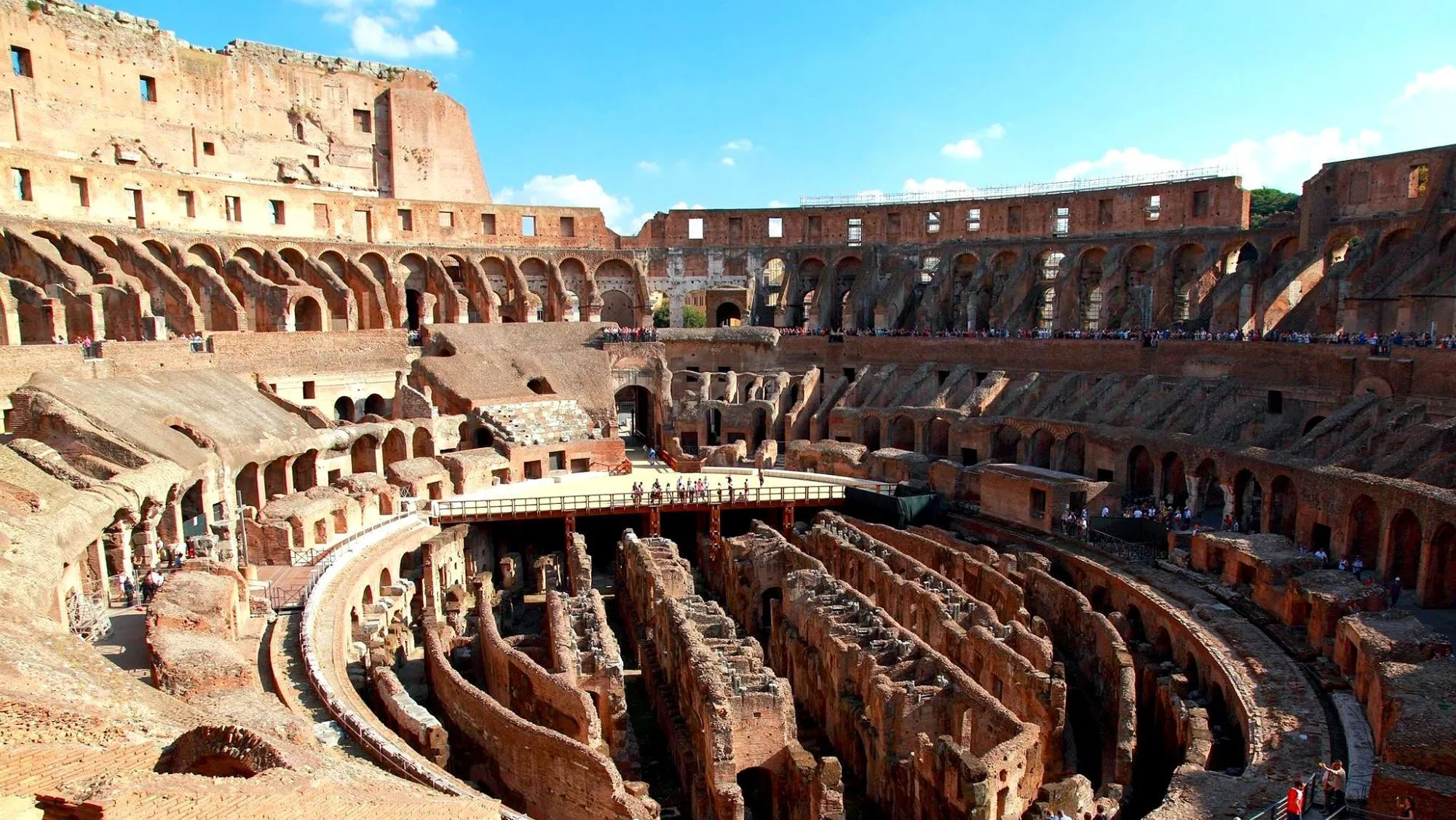 An expansive interior view of the ancient Colosseum in Rome, Italy, showcasing its immense scale and intricate tiered architectural ruins under a clear blue sky.