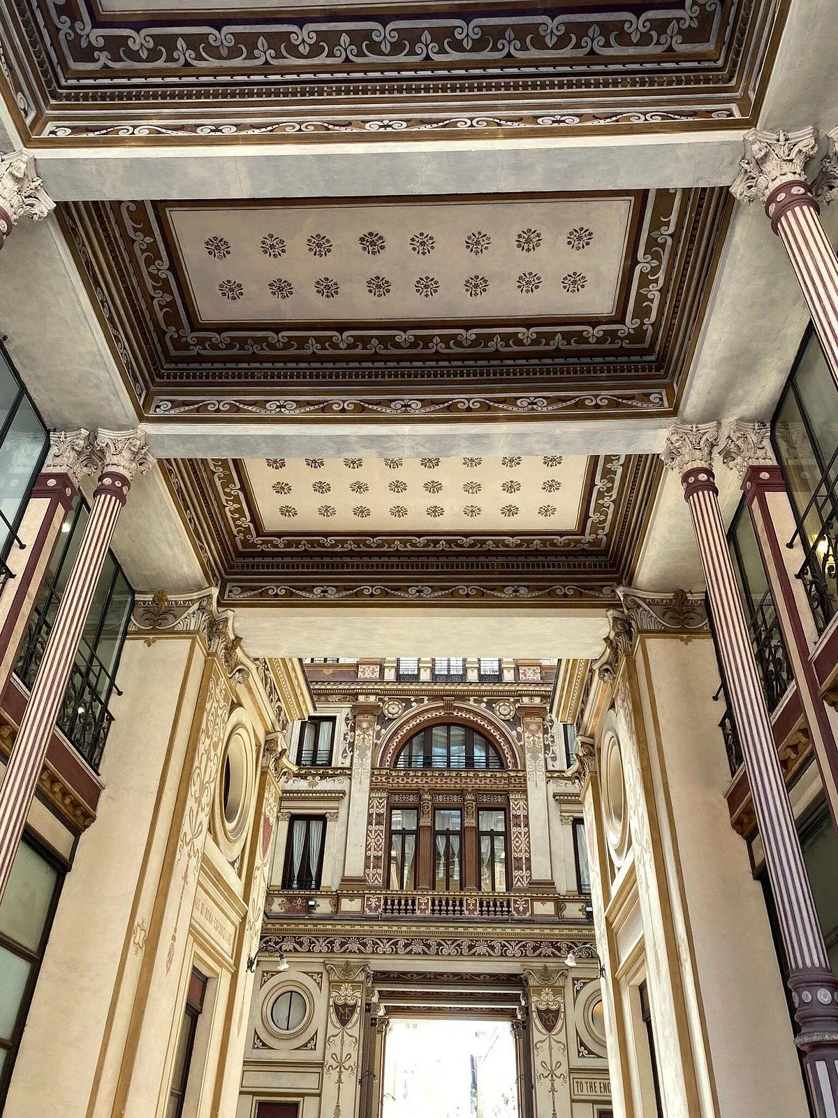 An interior shot of a grand, ornate architectural space, likely a historical hall or gallery, showcasing intricate ceiling patterns, decorative columns, and detailed facades with arched windows. The perspective emphasizes the depth and symmetry of the elaborate design.
