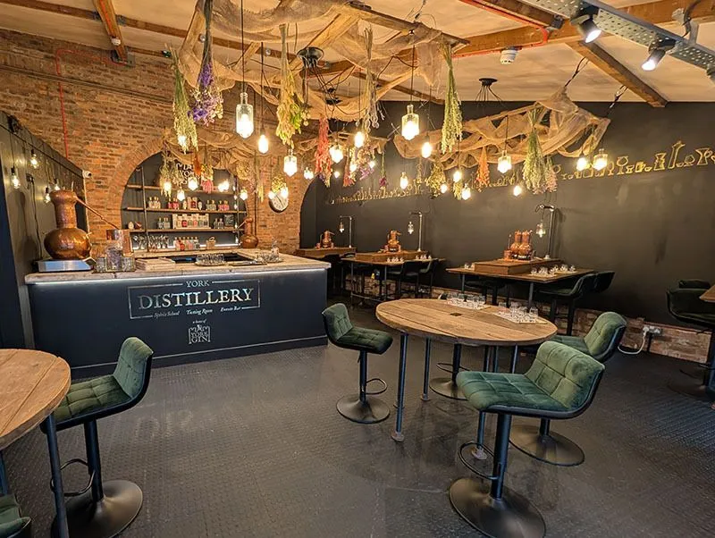 An interior shot of a distillery bar area, featuring a long dark bar with "YORK DISTILLERY" signage, several high-top tables with velvet green chairs, and atmospheric lighting from numerous hanging bulbs and decorative elements on the ceiling.