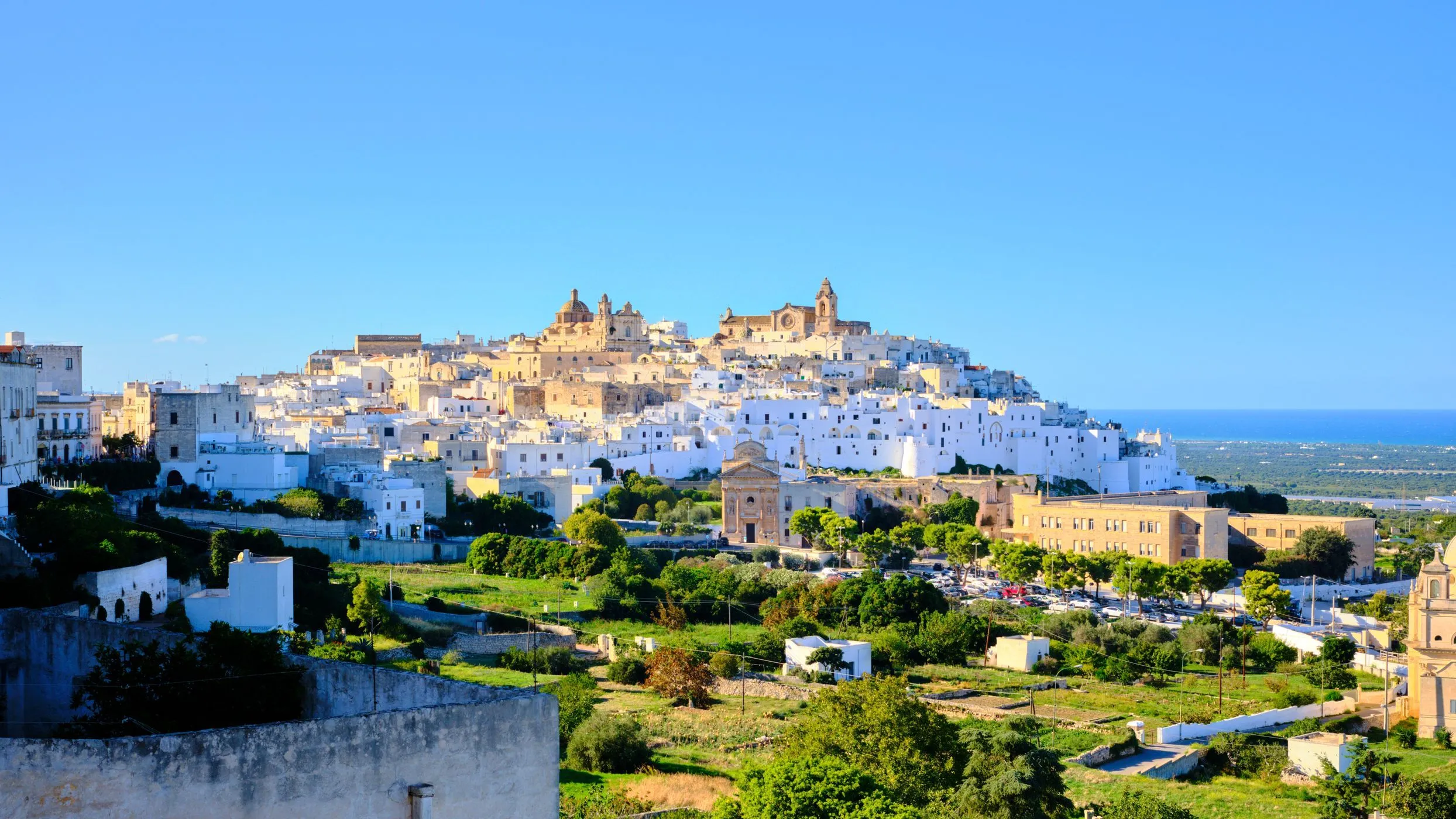 An editorial-style image showcasing a picturesque white-washed hill town under a clear blue sky, surrounded by green landscape. The architecture features distinctive domed buildings and intricate details.
