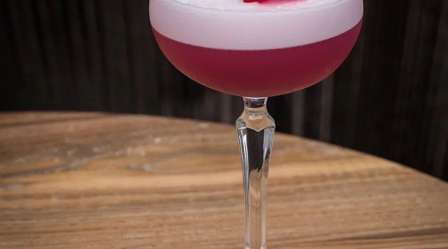 The image features a visually appealing cocktail in a stylish glass, exhibiting excellent composition with the drink as the central focus. The lighting is well-executed, highlighting the vibrant colors and textures of the beverage and its foamy top. The image is sharp, with all key elements in clear focus, contributing to a high level of photo-realism and accurate color representation. The drink appears full and professionally presented, set against a subtly blurred background with a clean wooden table.