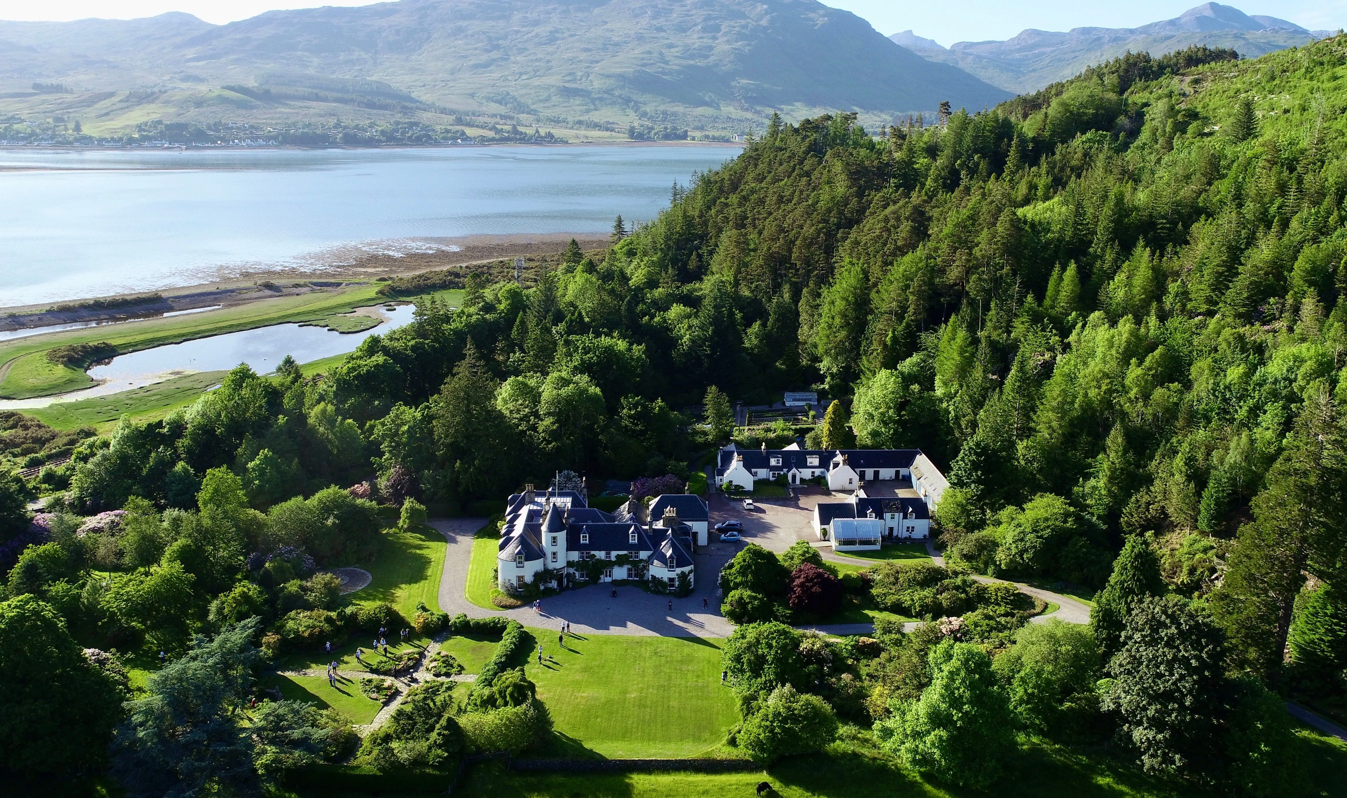 An aerial shot capturing a grand estate nestled in a lush green valley beside a large body of water and surrounded by forested hills and distant mountains.