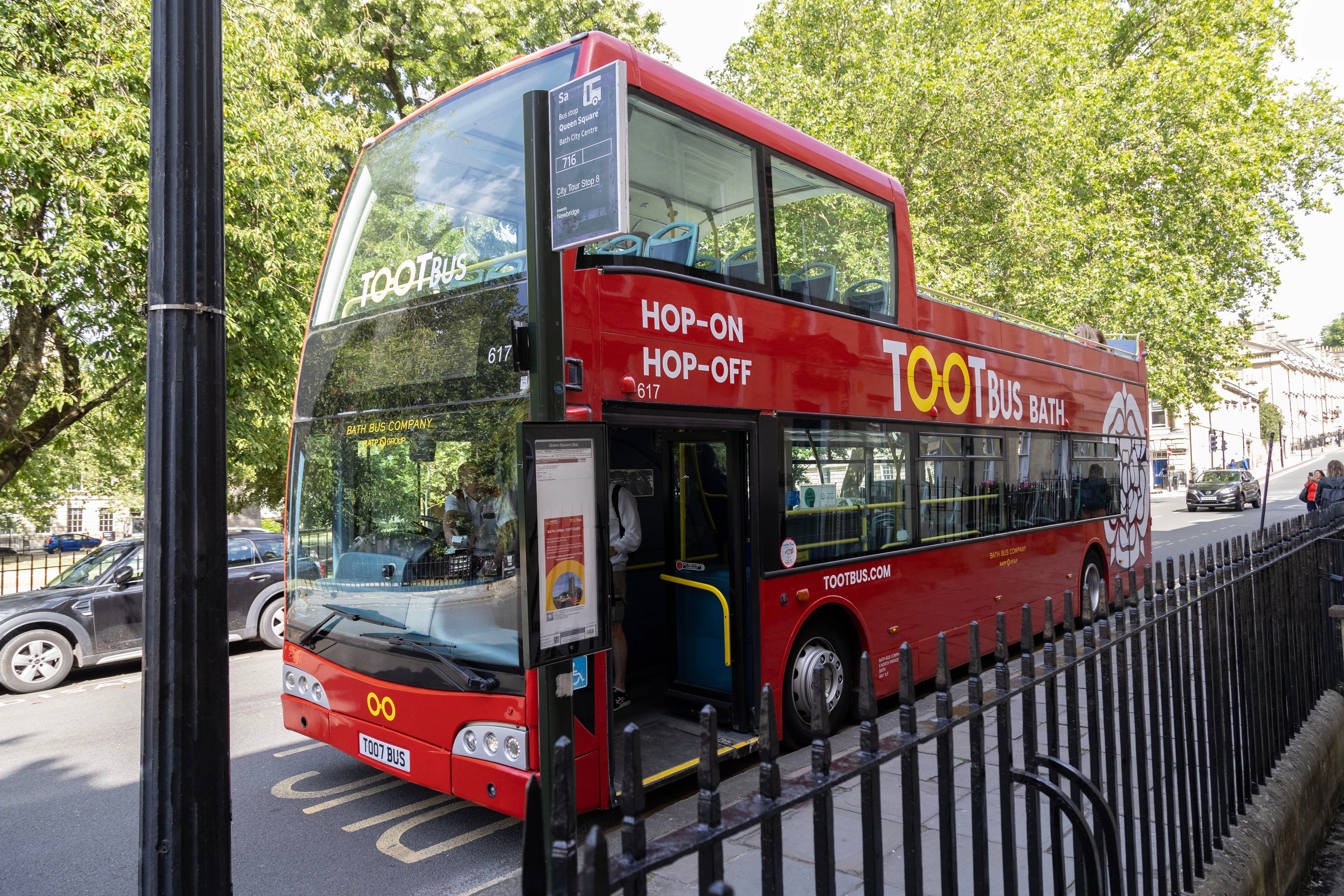 The image features a red double-decker 'Tootbus' tour bus, typical of a hop-on hop-off service, captured in what appears to be an urban setting with trees in the background. The bus is well-lit, sharp, and prominently displayed, showcasing its branding and details clearly. The composition effectively presents the bus in an editorial style, highlighting its key features without excessive clutter.