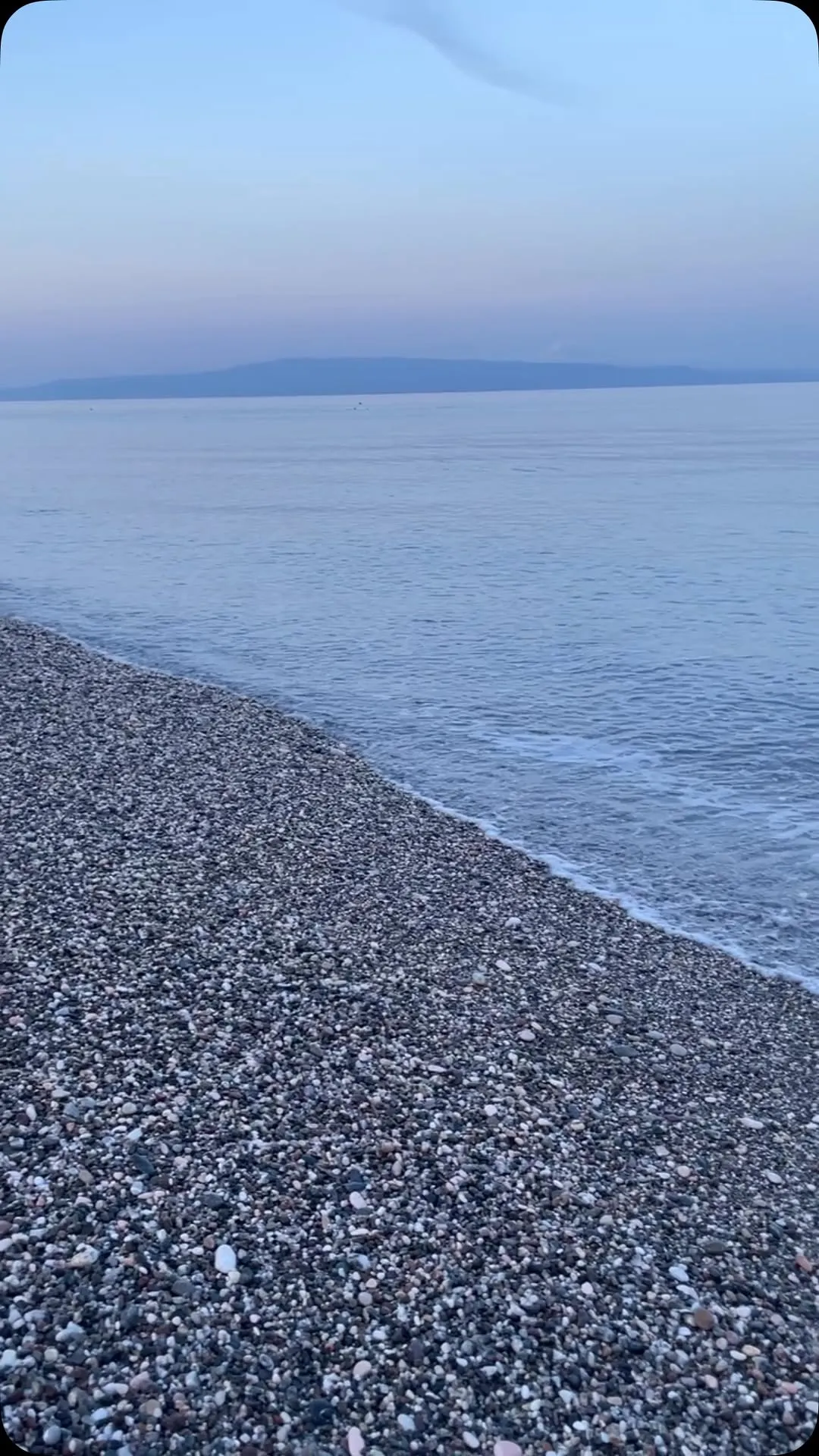 An outdoor shot of a pebble beach next to the sea with distant mountains under a muted sky.