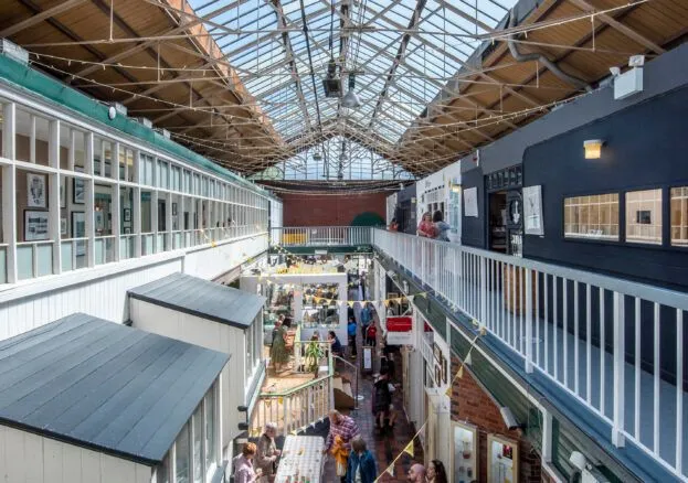 A well-composed and well-lit photograph capturing the vibrant atmosphere of an indoor market with a distinctive glass-roofed architectural design. The image is sharp, color-accurate, and highly photo-realistic, with all key elements in focus.