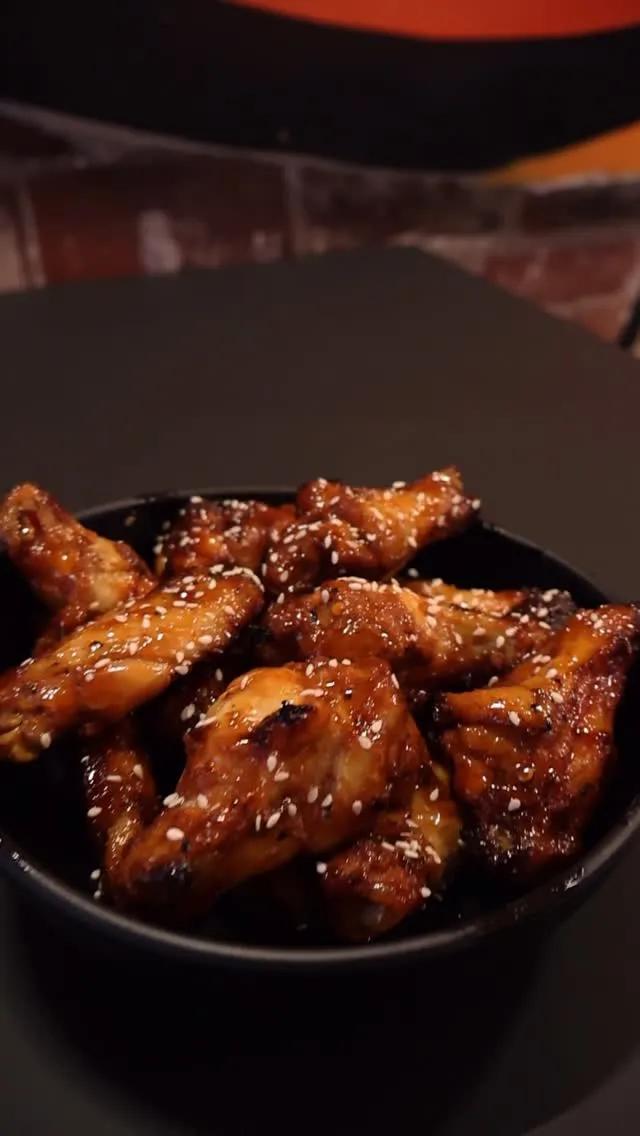The image features a close-up, well-composed shot of glazed chicken wings with sesame seeds in a dark bowl. The main subject is sharp and in focus, showcasing the textures and appealing colors of the food. The lighting is good, contributing to the photo-realistic quality.
