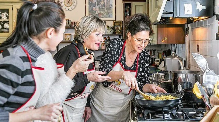An image capturing three women engaged in cooking within a kitchen setting, dressed in aprons. One woman is stirring a pan on the stove, while the others observe and interact. The image conveys a sense of communal activity and learning.