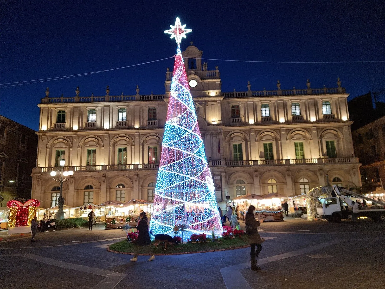 The image features a well-composed shot of a large, illuminated Christmas tree centered in a public square, with an ornate building providing an architectural backdrop. The tree's lights are vibrant and the building is nicely illuminated. The scene captures a festive atmosphere with market stalls and people, fitting an editorial style. The image is highly photo-realistic and the primary subjects, the tree and building, are sharp and in focus. The foreground, however, is a bit dark, which is common for night photography, and some transient elements like people are not perfectly sharp, though this does not detract from the main subject.