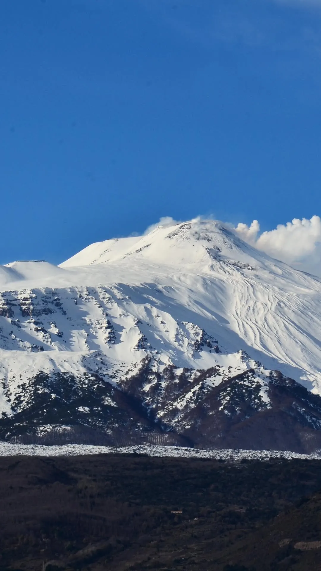 The image prominently features a majestic, snow-capped mountain under a clear blue sky. The mountain dominates the frame, showcasing its rugged, snow-covered slopes and a visible plume of steam or cloud near its peak, suggesting volcanic activity. The lighting is bright, highlighting the texture of the snow and creating distinct shadows that add depth to the terrain.