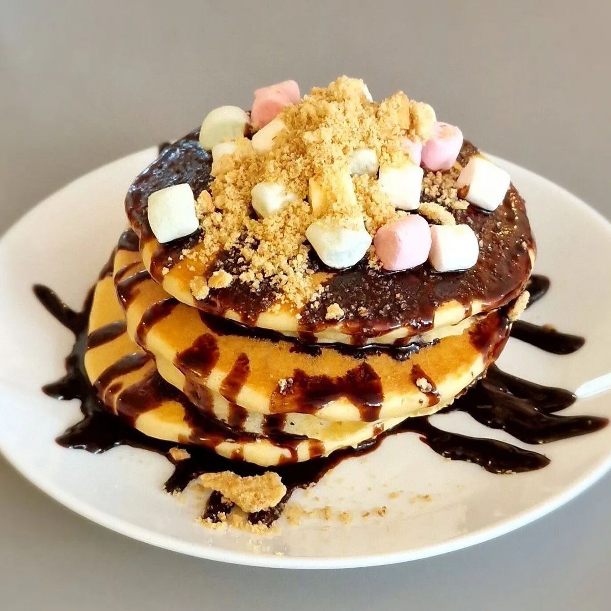 A well-composed and brightly lit close-up of a stack of pancakes topped with melted chocolate sauce, mini marshmallows, and graham cracker crumbs. The food is in sharp focus, showcasing appealing textures and vibrant, accurate colors, creating a highly photo-realistic and enticing image.