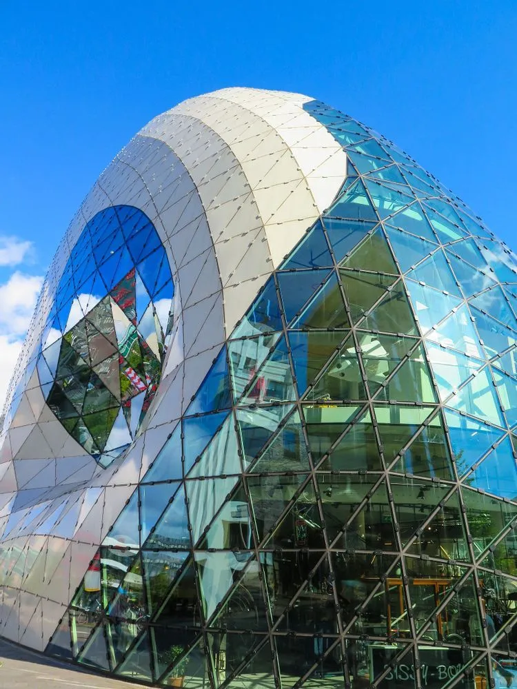 This image showcases a modern architectural structure with a striking facade composed of reflective glass and white panels. The composition is excellent, highlighting the unique curved design against a clear blue sky. The lighting is superb, illuminating the building evenly and enhancing its textures and reflections. The image is exceptionally sharp, with intricate details of the triangular panels and reflections clearly visible. Colors are vibrant and accurate, contributing to the overall photo-realistic quality. All key elements of the architecture are in perfect focus, making for a high-quality editorial image.