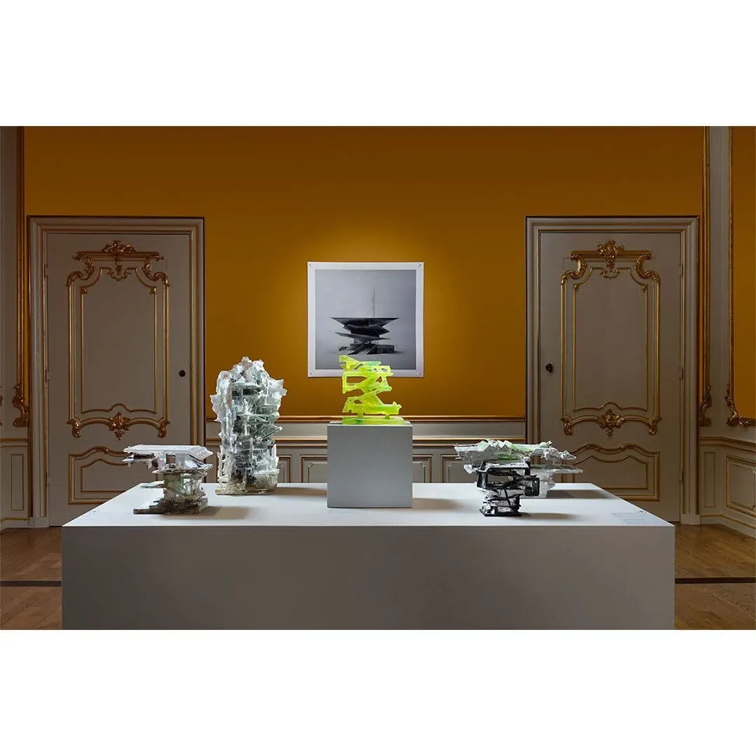An interior shot of an art exhibition featuring several abstract sculptures displayed on a white pedestal against a vibrant mustard yellow wall. The background includes ornate white doors and a framed artwork. The image showcases well-composed elements, effective lighting, and sharp details, with all key subjects in focus.