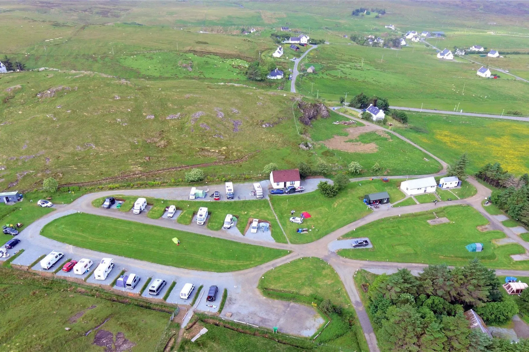 An aerial view of a green campsite with various caravans and buildings, nestled within a natural, undulating landscape under overcast skies.