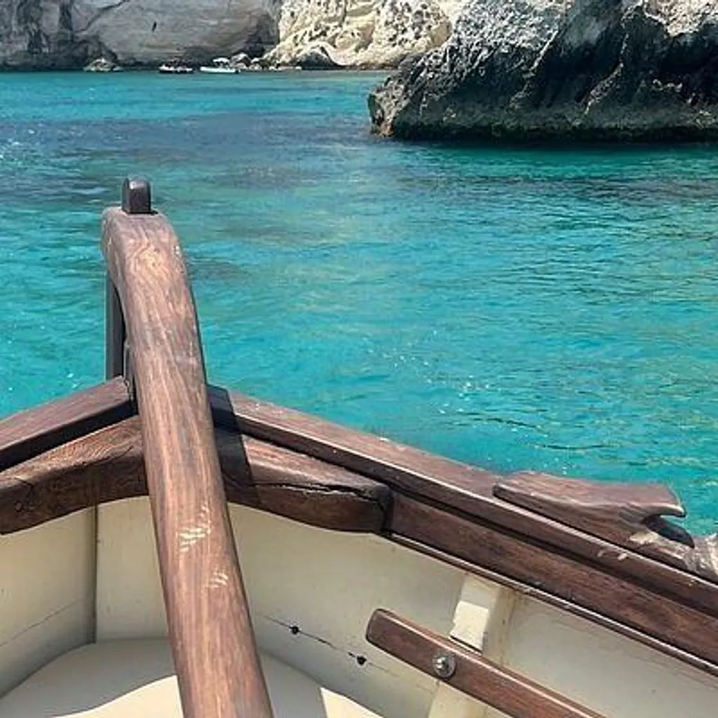 A view from the bow of a small wooden boat on calm, clear turquoise waters, approaching a rocky coastline with cliffs and sea caves under a bright sky.