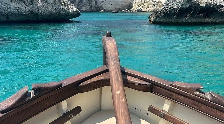 A view from the bow of a small wooden boat on calm, clear turquoise waters, approaching a rocky coastline with cliffs and sea caves under a bright sky.