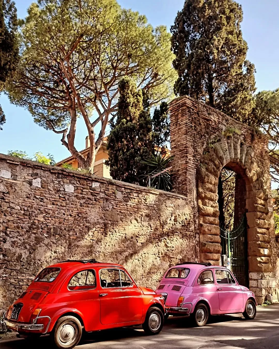An outdoor photograph showcasing two classic Fiat 500 cars, one red and one pink, parked on a street next to an old stone wall and a large brick archway. Trees are visible in the background under a clear sky.