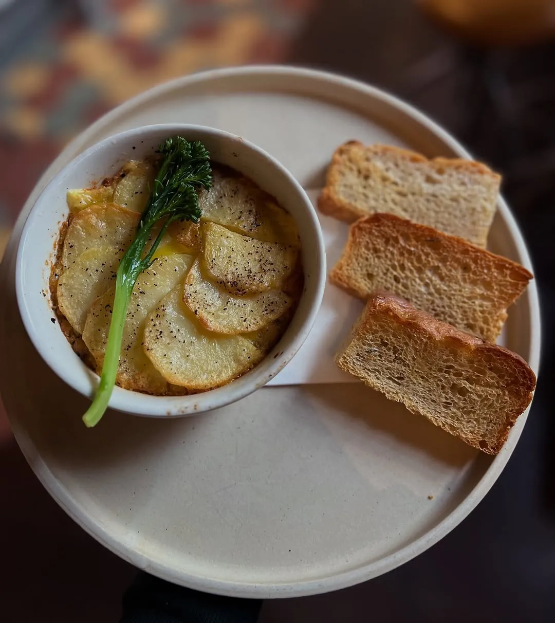 A close-up, top-down view of a savory potato dish, possibly a gratin or dauphinoise, served in a white bowl with a sprig of broccoli. Three slices of toasted bread accompany the dish on a light, round plate. The background is softly blurred.
