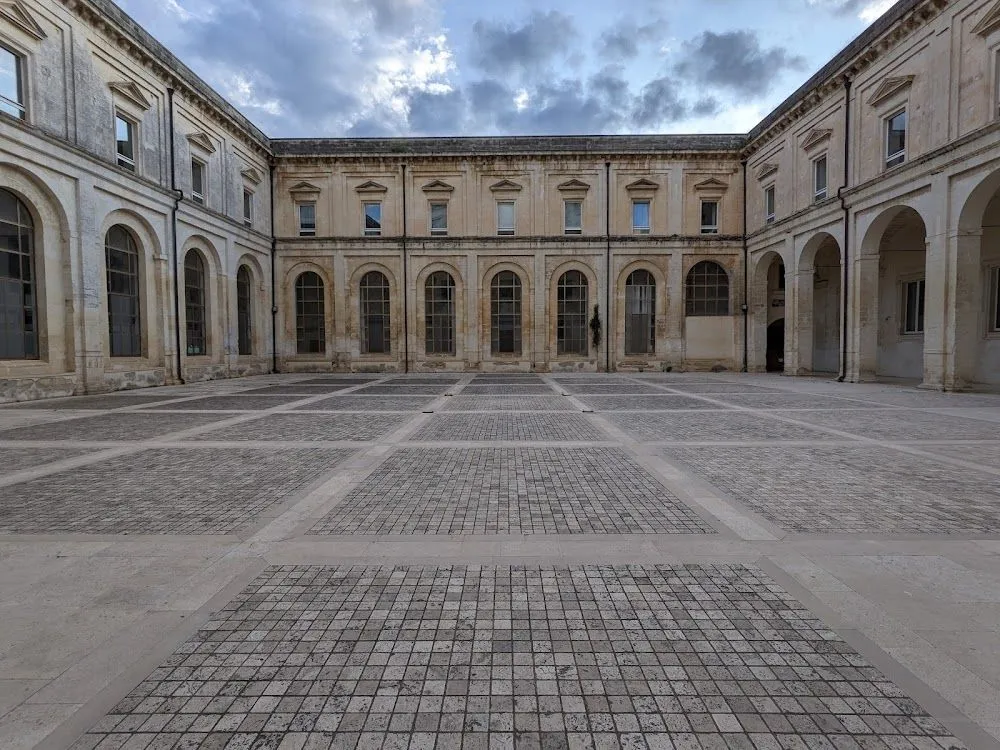 An exterior shot of a large, U-shaped historical building surrounding a paved courtyard under an overcast sky.