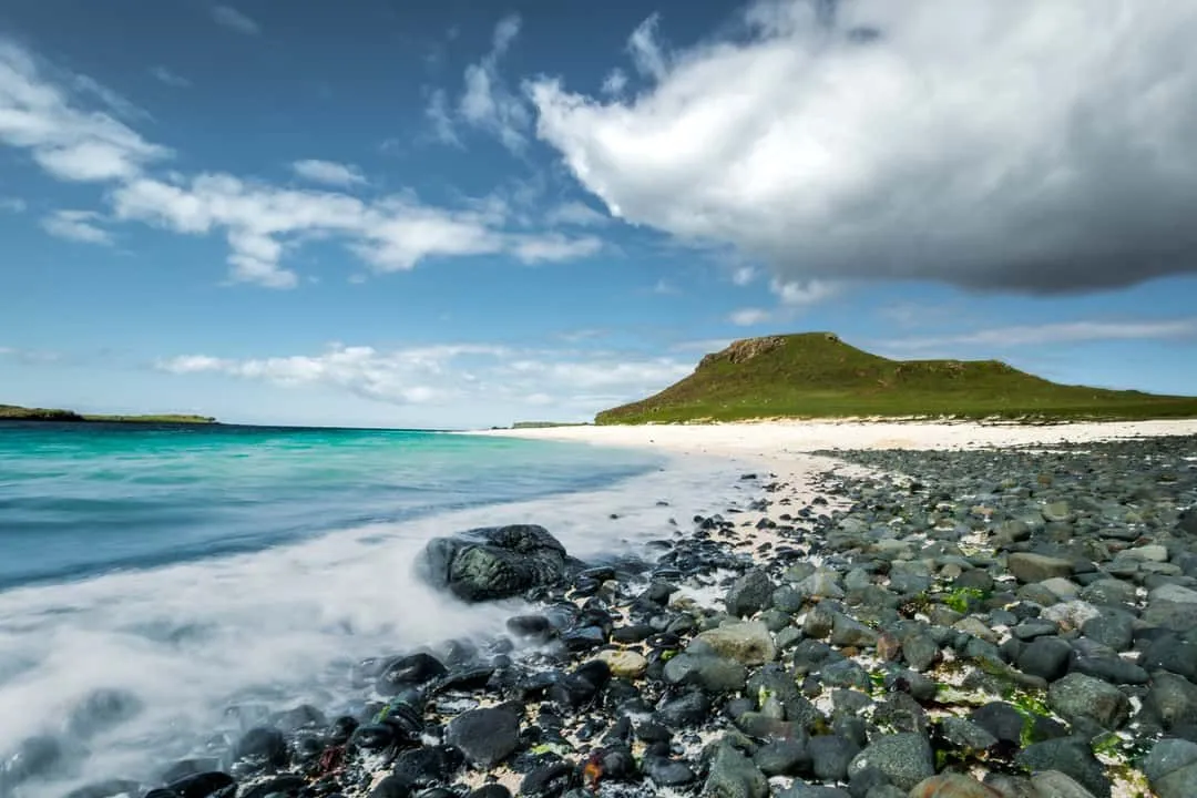 An editorial quality image of a serene beach landscape, featuring a rocky foreground leading to a white sandy beach, turquoise waters, and a green hill under a blue sky with clouds.