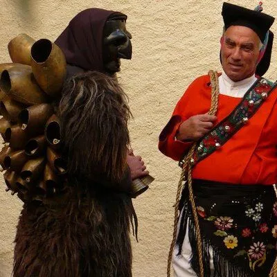 An image depicting two individuals in traditional attire, likely at a cultural event or festival. One figure wears a dark mask, a hooded garment, and a costume adorned with large bells and fur. The other, an older man, is dressed in a vibrant red jacket, a decorated sash with bells, and a black skirt with floral embroidery, holding a rope. They are set against a plain, light-colored wall.