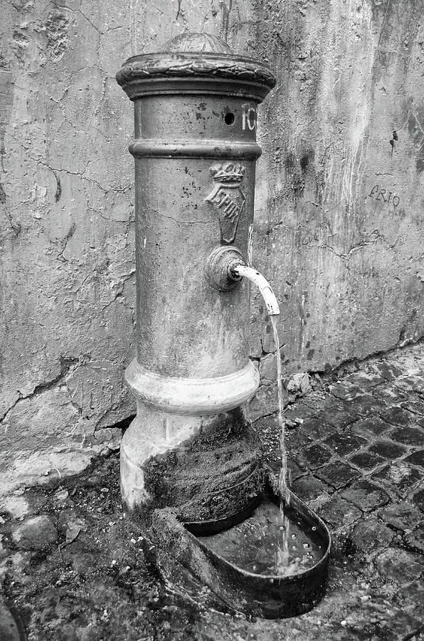 A close-up, black and white photograph of an old, weathered public water fountain (Nasoni-style) attached to a cracked, textured wall. Water flows continuously from the spout into a small basin below.