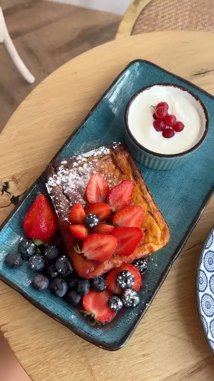 An overhead shot of French toast generously topped with fresh strawberries, blueberries, and powdered sugar, presented on a blue rectangular plate. A small side bowl contains yogurt or cream garnished with red currants.