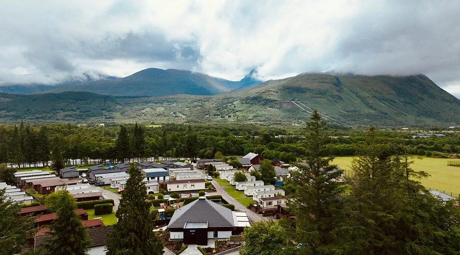 An elevated view of a caravan park situated in a valley, surrounded by lush green mountains under a cloudy sky. The image features numerous static caravans arranged along roads, with a central building and evergreen trees in the foreground.
