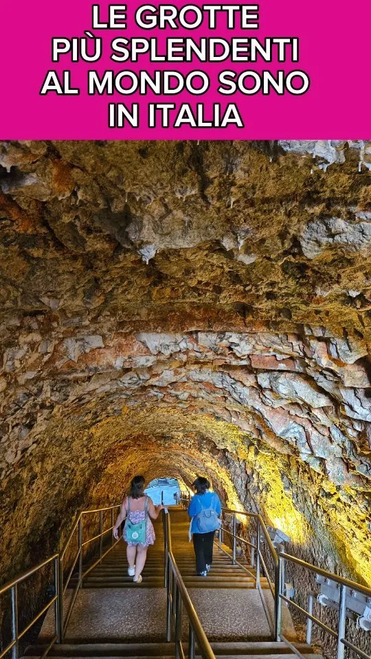 The image captures a well-lit tunnel or cave-like structure with rough, textured rock walls, creating a sense of depth and perspective. Warm artificial lighting illuminates the scene, highlighting the details of the rock formations. Two individuals are visible at the far end of the tunnel, providing a sense of scale and human element.