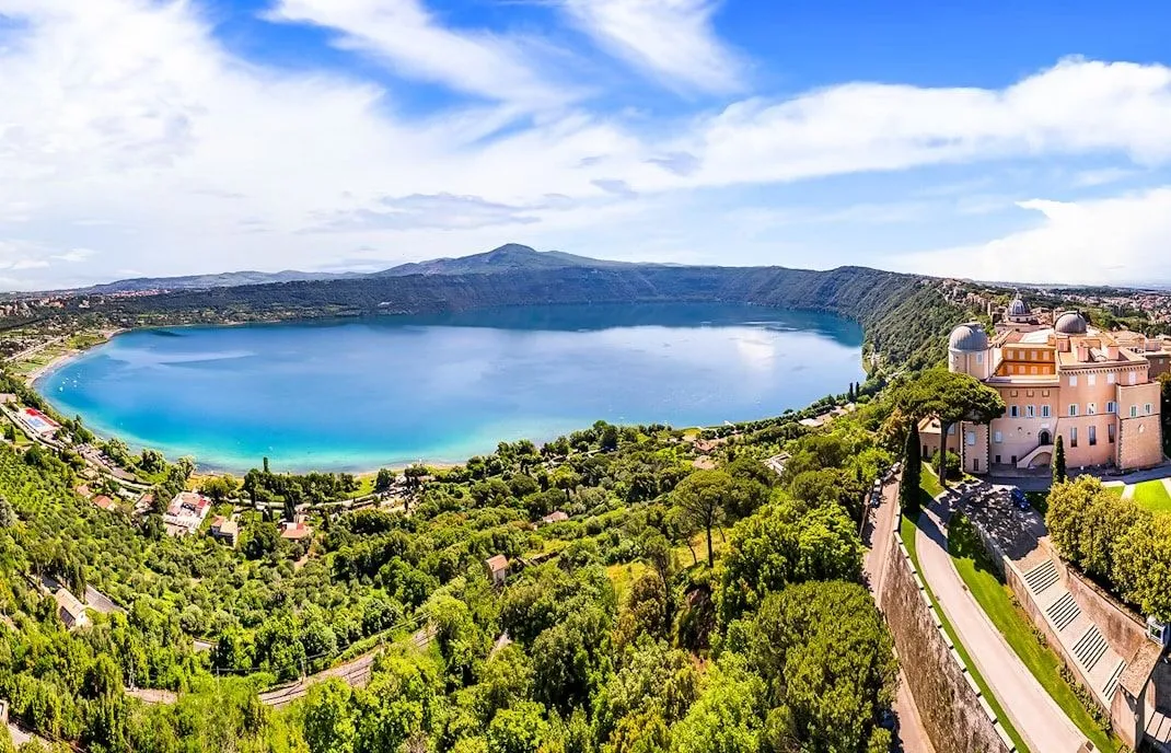 An expansive, high-angle view of a serene lake nestled within a verdant, mountainous landscape under a bright blue sky with scattered clouds. Lush green foliage covers the surrounding hills, with a winding road and some structures visible on the right side.