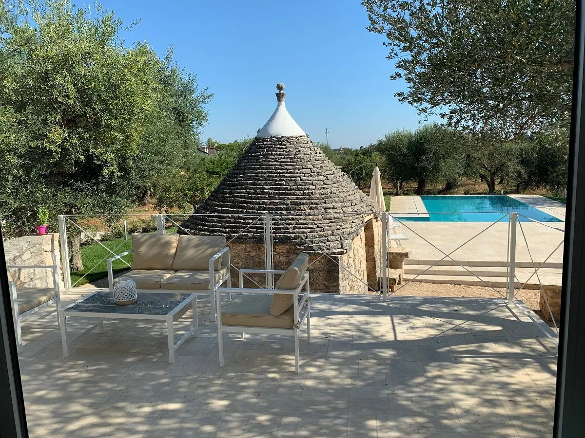 The image is sharp and photo-realistic, accurately capturing the colors of the outdoor scene under bright sunlight. The key elements, including the Trullo and the patio furniture, are clearly in focus. The composition effectively frames the Trullo and integrates the pool area into the shot.