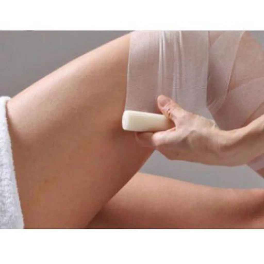 An editorial image showing a person's hand holding a wax strip and applying it to their leg for hair removal. The image focuses on the process of waxing.