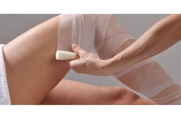 An editorial image showing a person's hand holding a wax strip and applying it to their leg for hair removal. The image focuses on the process of waxing.